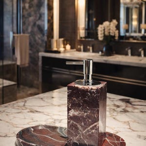 May include: A marble soap dispenser with a matching tray. The dispenser is a rectangular block of red and white marble with a silver pump. The tray is oval-shaped and made of the same marble. The set is on a marble countertop.