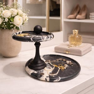 May include: A two-tiered jewelry stand with a black and white marble design. The stand has a black base and stem, with a small black sphere on top. Gold necklaces and bracelets are displayed on the stand.