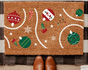 May include: Brown doormat with a Christmas ornament design. The mat features red, green, and white ornaments, candy canes, snowflakes, and strings of lights. The design is set against a brown background.