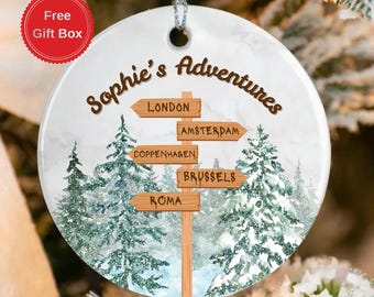 May include: A white ceramic ornament with the text "Sophie's Adventures" and a signpost pointing to London, Amsterdam, Copenhagen, Brussels, and Rome. The ornament features a winter forest scene with snow-covered trees and a "Free Gift Box" sticker.