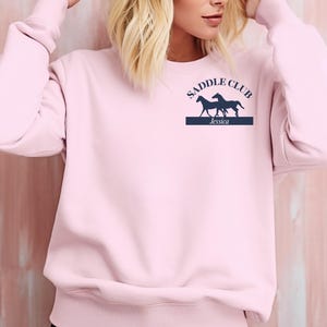 May include: Pale pink sweatshirt featuring the words "SADDLE CLUB" above two dark blue horse silhouettes. Underneath the horses is a dark blue rectangle with the name "Jessica" in white text.