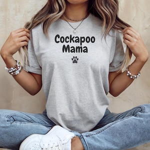 May include: A light gray t-shirt with the words "Cockapoo Mama" in black, along with a paw print graphic. The shirt is made of a soft material and has rolled-up sleeves. The shirt is a casual style.