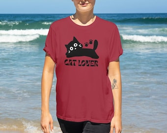 May include: A maroon t-shirt with a black cat graphic and the text "CAT LOVER". The cat is lying down with its paws up. The background is a beach with blue water and waves.