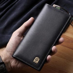 May include: A black leather wallet with a gold emblem. The wallet is rectangular and appears to be closed. The gold emblem is on the lower right corner. The wallet is held in a hand.