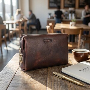 May include: A brown leather clutch with a zipper closure and a gold-colored lock. The clutch is on a wooden table next to a cup of coffee, a notebook, and a pen. The clutch has the word "COLLOGS" embossed on it.
