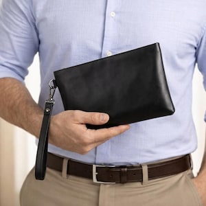 May include: A black leather clutch with a wrist strap is held by a person wearing a light blue button-down shirt and khaki pants. The clutch has a rectangular shape and a silver-colored clasp.