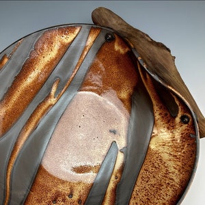 Può includere: Un piatto da portata in ceramica con una glassa marrone e nera. Il piatto ha una forma unica con un bordo curvo e un manico in legno.