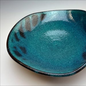 Può includere: Un piatto da portata in ceramica blu-verde con un bordo ondulato e una glassa marrone scuro. Il piatto ha una texture punteggiata e un sottile motivo di linee marroni scure.