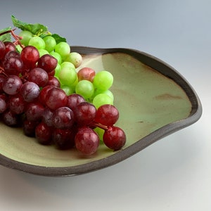 Può includere: Una ciotola di ceramica verde con un bordo ondulato, piena di uva rossa e verde. La ciotola ha un bordo marrone scuro.
