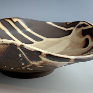 Può includere: Una ciotola da portata in ceramica con un bordo ondulato e un design astratto marrone e bianco. La ciotola ha una base leggermente rialzata.