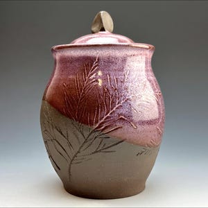 Puede incluir: Un tarro de cerámica con tapa, decorado con un esmalte rosa y marrón y un diseño de hojas. El tarro tiene un cuerpo redondeado y una tapa pequeña y redonda.
