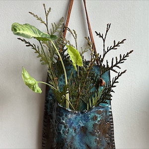 Op de afbeelding: Een tealblauwe keramische wandpot met een bruine leren hangriem. De pot is gevuld met groene, bladerige planten.