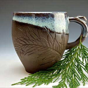 May include: A brown ceramic mug with a white glaze and a fern design. The mug has a unique handle and is sitting on a white surface with a green sprig of cedar.