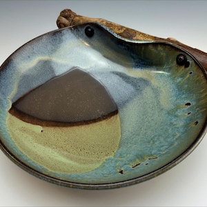 Può includere: Una ciotola in ceramica fatta a mano con un design unico. La ciotola presenta un centro marrone scuro, circondato da una glassa verde chiaro e blu. Un pezzo di legno è attaccato al bordo, aggiungendo un elemento naturale.
