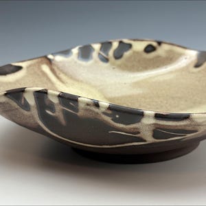 Può includere: Una ciotola in ceramica dalla forma organica unica. La ciotola presenta un interno color crema con motivi astratti marrone scuro e neri lungo il bordo. L'esterno è di colore marrone scuro e poggia su una piccola base circolare.