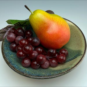 Può includere: Una ciotola di ceramica con bordo verde e marrone contiene un grappolo di uva rossa, una pera gialla e arancione e una foglia verde. La pera è posizionata sul lato destro della ciotola, con l'uva a sinistra.