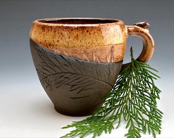 May include: A handmade ceramic mug with a brown and tan glaze. The mug has a unique design with a fern pattern on the bottom half. The mug is sitting on a white surface with a green sprig of cedar.