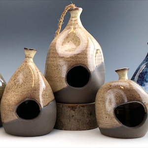 Puede incluir: Cinco casas para pájaros de cerámica en varios colores y estilos. Las casas para pájaros están hechas de arcilla y tienen un aspecto rústico y hecho a mano. Todas son de diferentes tamaños y formas, pero todas tienen un agujero en la parte delantera para que los pájaros puedan entrar.