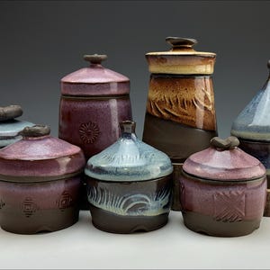 Può includere: Una collezione di sette vasi in ceramica fatti a mano con vari colori e motivi. I vasi sono di diverse dimensioni e forme, alcuni con coperchi. I vasi sono disposti in fila su una superficie di legno.