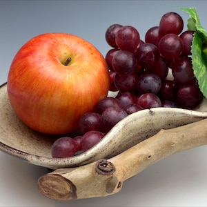 Puede incluir: Un plato de cerámica con un asa de madera natural contiene una manzana roja y un racimo de uvas rojas. El plato tiene un interior beige y un borde marrón oscuro. Una hoja verde acentúa las uvas, creando una naturaleza muerta.