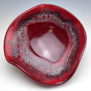 Puede incluir: Un cuenco de cerámica hecho a mano, de forma irregular. El cuenco es de color rojo intenso con un acabado brillante. El borde presenta un patrón texturizado y moteado en blanco y negro, contrastando con el rojo.