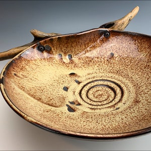 Può includere: Una grande ciotola in ceramica con una glassa marrone e gialla. La ciotola ha una forma unica e organica ed è decorata con un motivo a spirale al centro. Un pezzo di legno di deriva è attaccato al bordo della ciotola.