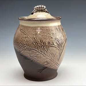 Puede incluir: Un tarro de cerámica hecho a mano con tapa. El tarro es una mezcla de marrón y beige con una superficie texturizada y un diseño detallado de hojas. La tapa tiene un asa texturizada. El tarro es un recipiente decorativo.