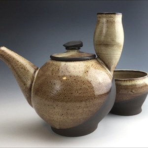 Può includere: Un set da tè in ceramica fatto a mano. La teiera è marrone con coperchio e beccuccio neri. La tazza è marrone e bianca. Il set viene probabilmente utilizzato per il tè o altre bevande calde.