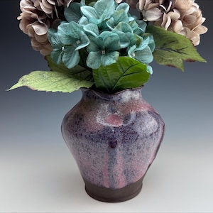 May include: A ceramic vase with a dark purple and pink speckled glaze, holding faux blue and tan hydrangeas. The vase has a dark brown base and a ruffled rim, creating a decorative floral arrangement.