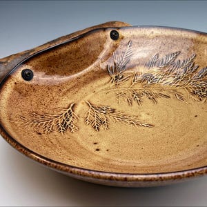 Può includere: Un grande piatto da portata in ceramica marrone con un manico in legno. Il piatto ha una superficie testurizzata con un disegno di foglie di felce. Il manico è fissato al piatto con due viti in metallo.