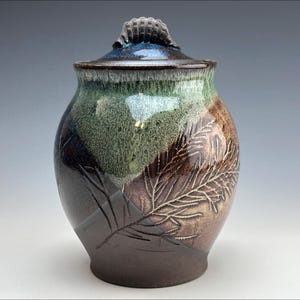 Puede incluir: Un tarro de cerámica con tapa, con un diseño de hojas. El tarro tiene una base marrón oscuro, que transiciona a tonos verdes y azules hacia la parte superior. La tapa tiene un asa texturizada. El tarro mide aproximadamente 20 cm de alto.