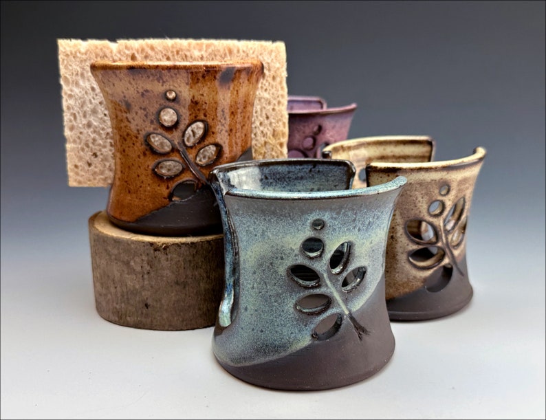 May include: Four ceramic sponge holders with a leaf design. The holders are in various shades of brown, green, and purple. The holders are made of pottery and have a rustic look.
