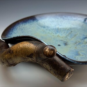 Può includere: Un piatto da portata in ceramica blu e marrone con un manico in legno. Il piatto ha una forma organica unica e una glassa macchiettata.