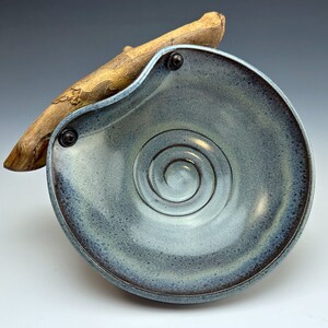 Può includere: Un piatto in ceramica blu con un design a spirale e un accento in legno. Il piatto ha una glassa testurizzata e un bordo leggermente irregolare.