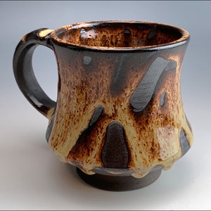May include: A handmade ceramic mug with a unique, textured glaze. The mug features a dark brown base and handle, with a blend of tan and brown tones on the body. Darker brown accents create a striking contrast. The mug is designed for hot beverages.