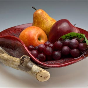 Può includere: Una ciotola per la frutta in ceramica rossa con un manico in legno naturale, piena di uva rossa, una mela rossa e una pera gialla.