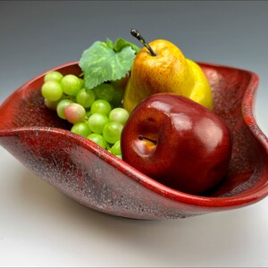 Può includere: Una ciotola di frutta in ceramica rossa con un bordo ondulato, piena di uva verde, una mela rossa e una pera gialla.