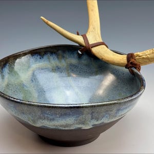 Può includere: Una ciotola in ceramica fatta a mano con una glassa blu e verde, con una base marrone scuro. Un corno di cervo è attaccato alla ciotola con cinghie di cuoio marrone, aggiungendo un'estetica rustica e naturale. Ideale per l'arredamento della casa.