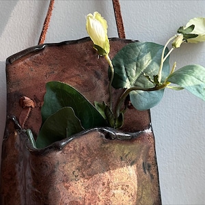 Puede incluir: Un bolsillo de pared artesanal, de color cobre, con borde oscuro y correa de cuero. Contiene hojas verdes y flores amarillas pálidas. La pieza tiene un aspecto rústico y texturizado, ideal para plantas pequeñas.