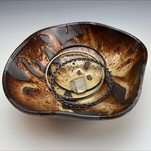Peut inclure: Un plat en céramique brun et jaune avec un bord ondulé. Le plat a un glaçage moucheté et est décoré de tourbillons bruns et noirs. Deux bracelets en argent et un pendentif en argent sont posés dans le plat.