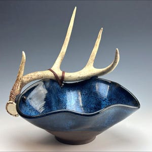 May include: A handcrafted ceramic bowl with a deep blue glaze and a brown base. A natural antler is attached to the bowl with a brown leather strap. The bowl has a unique, wavy rim.
