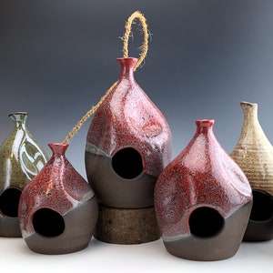 May include: A collection of six ceramic birdhouses in various colors and patterns. The birdhouses are all different shapes and sizes, and some have a hole in the front for birds to enter. The birdhouses are arranged in a row on a table.