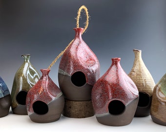 May include: A collection of six ceramic birdhouses in various colors and patterns. The birdhouses are all different shapes and sizes, and some have a hole in the front for birds to enter. The birdhouses are arranged in a row on a table.
