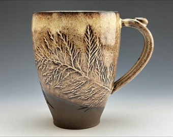 Könnte beinhalten: Eine braune und cremefarbene Keramiktasse mit einem Farnmuster. Die Tasse hat einen breiten Boden und einen abgerundeten Griff.