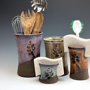 May include: Set of three ceramic kitchen utensil holders with a leaf design. The holders are in shades of brown, purple, and blue. The holders are designed to hold kitchen utensils, sponges, and brushes.