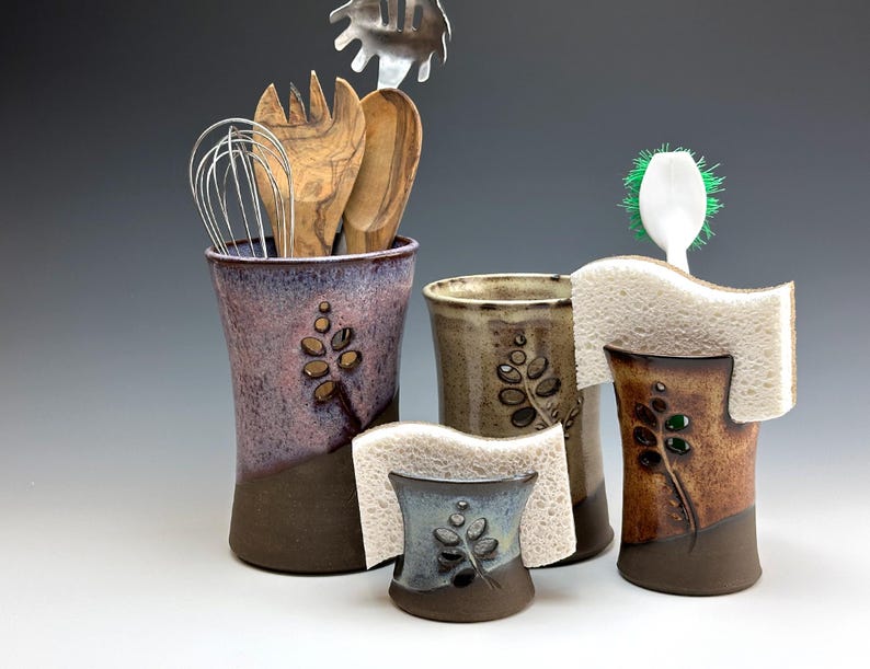 May include: Set of three ceramic kitchen utensil holders with a leaf design. The holders are in shades of brown, purple, and blue. The holders are designed to hold kitchen utensils, sponges, and brushes.