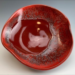 Può includere: Una ciotola in ceramica rossa con una glassa craquelé bianca. La ciotola ha una forma ondulata, organica e un interno profondo e arrotondato.