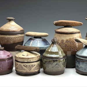 Puede incluir: Una colección de pequeños botes de cerámica con tapa, con diversos diseños y colores. Las tapas son de madera y tienen un acabado natural. Los botes están dispuestos en fila sobre una superficie blanca.