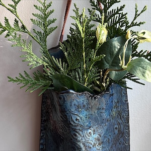 May include: A blue ceramic wall vase with a textured surface and a decorative pattern. The vase holds a variety of greenery, including evergreen branches and a few delicate flowers. A brown leather strap is attached for hanging.