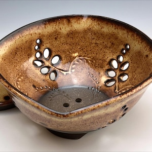May include: A brown ceramic colander with a square shape and a decorative leaf design. The colander has small holes throughout the bottom and sides. It is sitting on a matching brown ceramic plate.